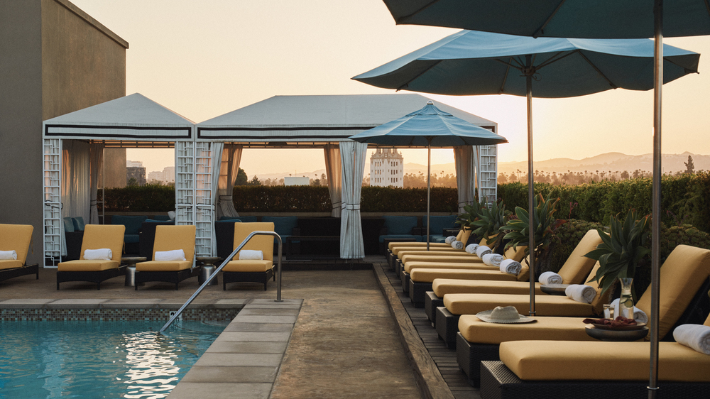 L'Ermitage Beverly Hills pool area with views of surrounding Los Angeles during dusk with poolside canary yellow lounge chairs and covered cabanas.