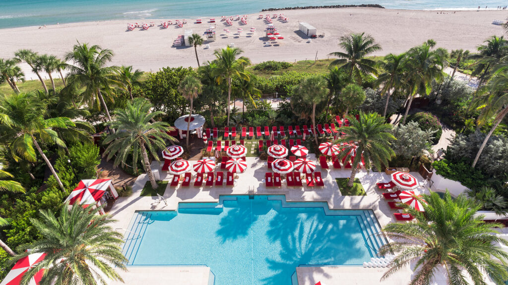 Aerial view of Faena pool area with poolside red lounge chairs with aesthetically pleasing red and white umbrellas with views of the beach with matching umbrellas in the sand. 