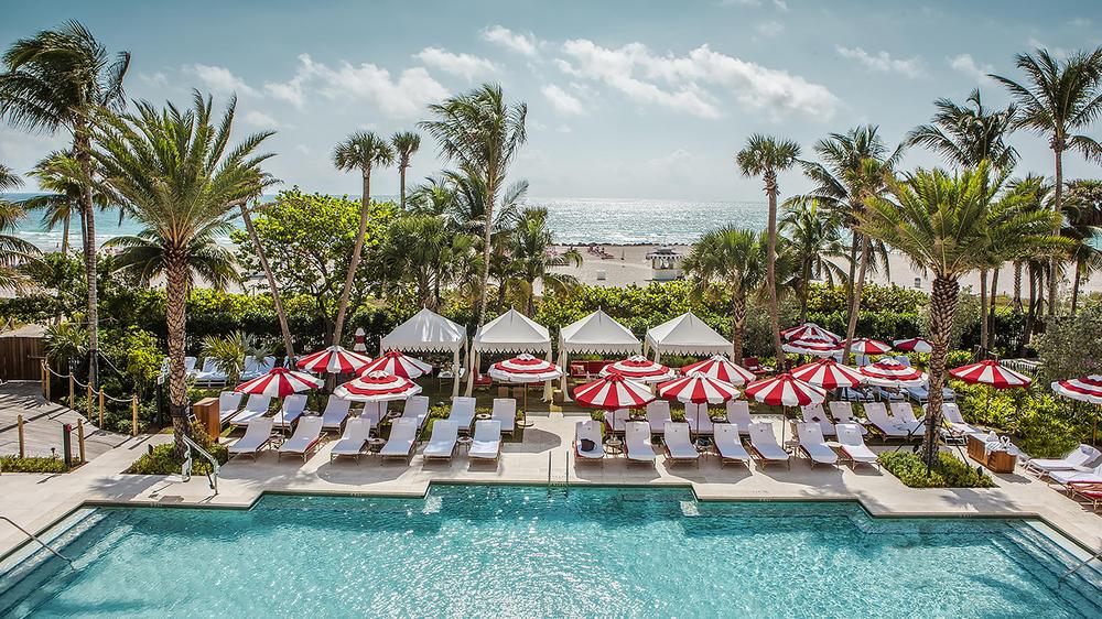 Faena Hotel Miami Beach pool area with poolside white lounge chairs and vibrant red and white umbrellas with a garden separating the pool area and hotel beach during a sunny day. 