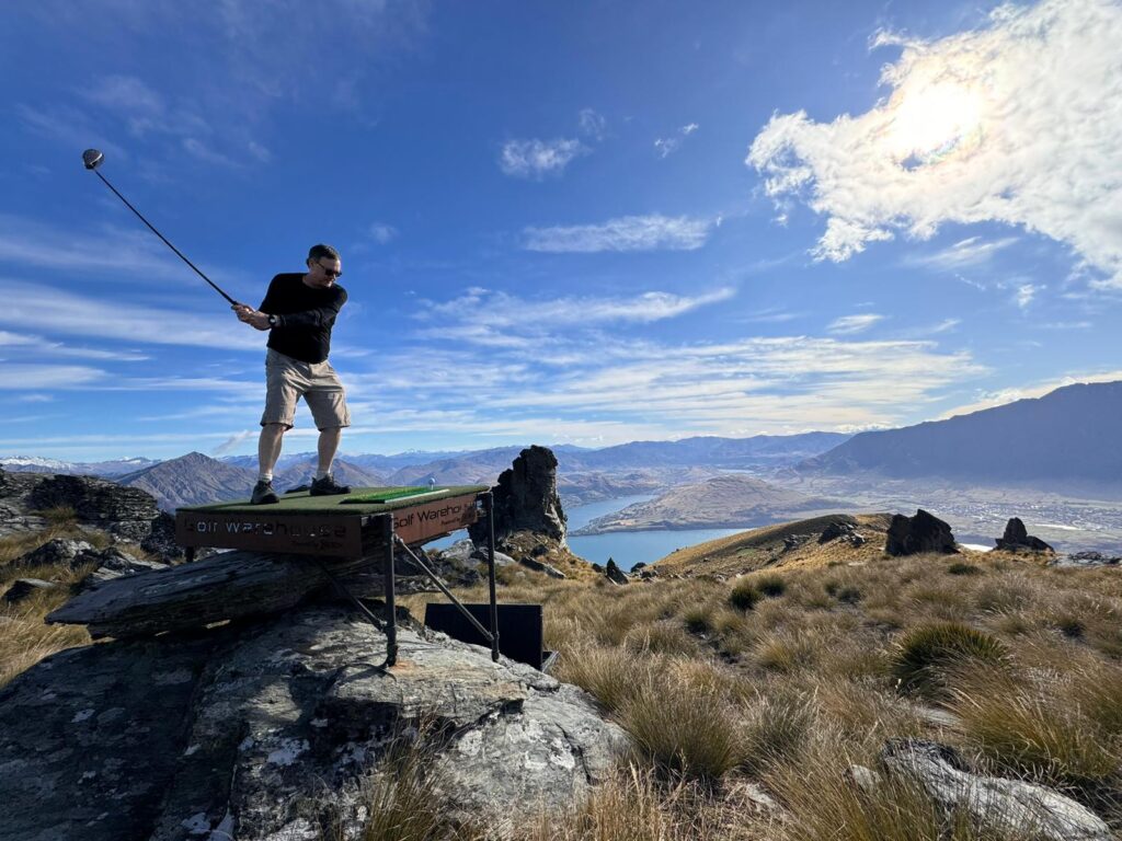 A Inspirato member swinging a golf club on a raise platform in front of a mountain and lake vista