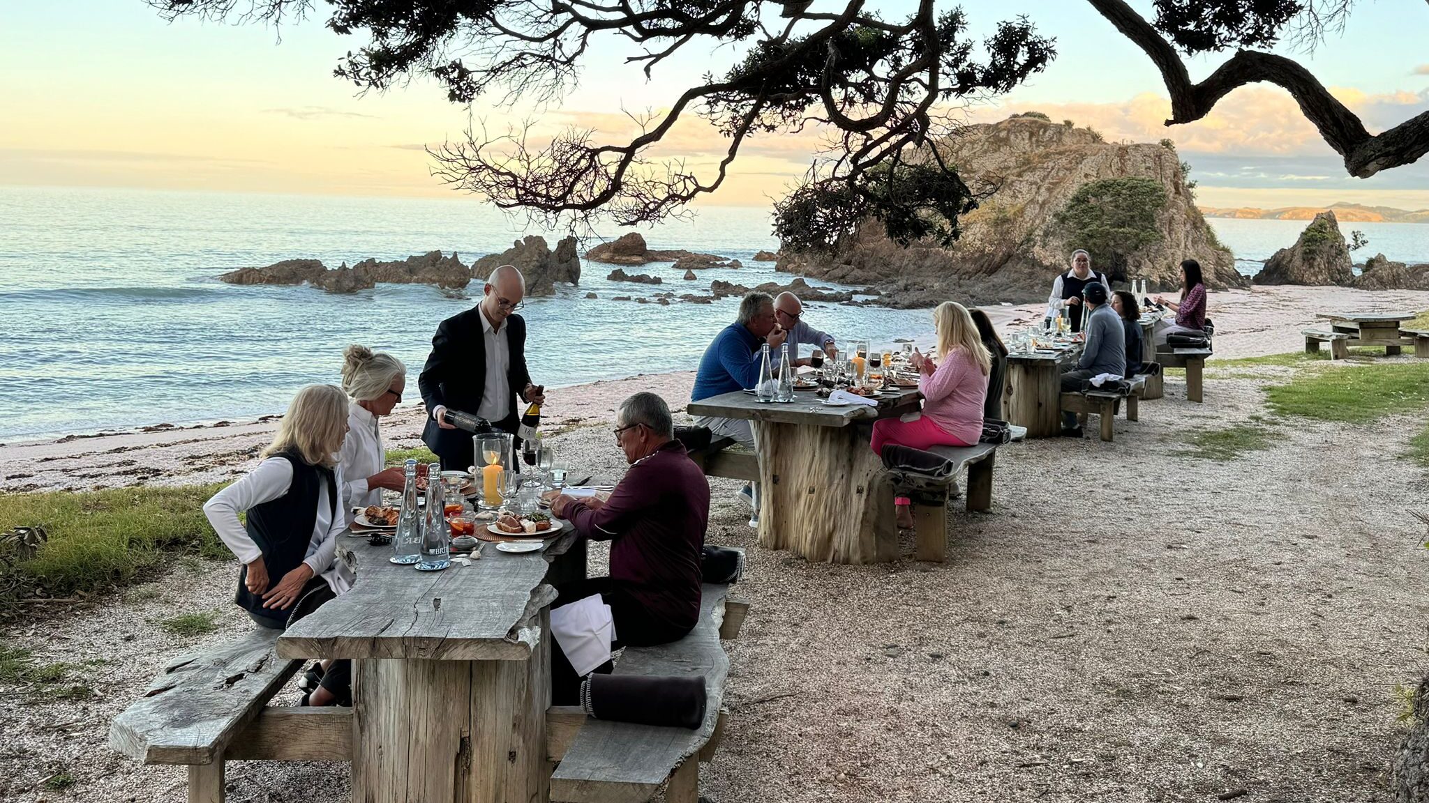 Inspirato members sitting a wooden tables on the beach in New Zealand enjoying a curated dinner