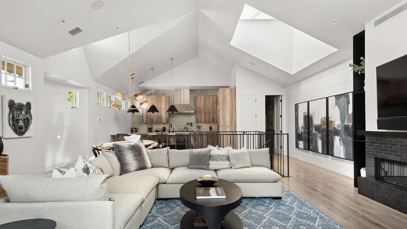 Buckhorn living area with vaulted white ceiling and a large white sectional couch in front of a wall-mounted flat screen tv hanging above a fireplace with kitchen area and stairs leading back down to ground level. 