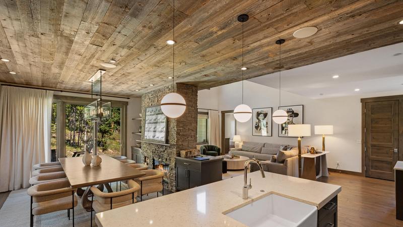 Flatiron dining and living area with large wooden table that seats ten with modern light fixtures hanging above the kitchen island and large stone fireplace that divides the dining room and living area. 