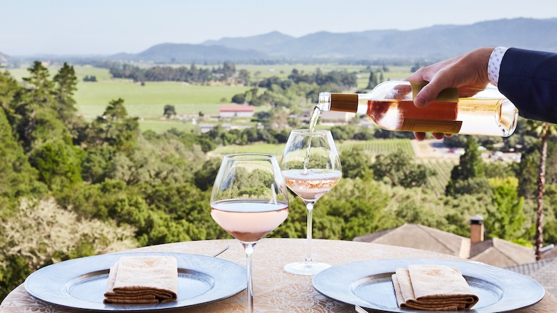 Set up of dining table with someone pouring wine into two glasses with views of Napa Valley.