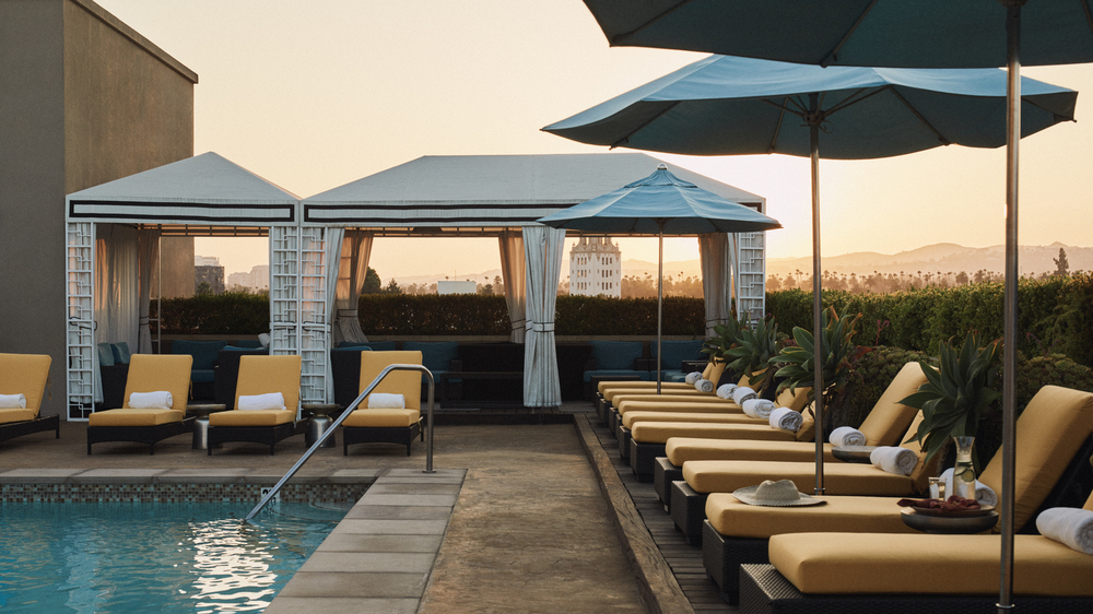 L'Ermitage pool area with poolside canary lounge chairs with blue umbrellas with white cabana with views of surrounding Los Angeles during dusk. 