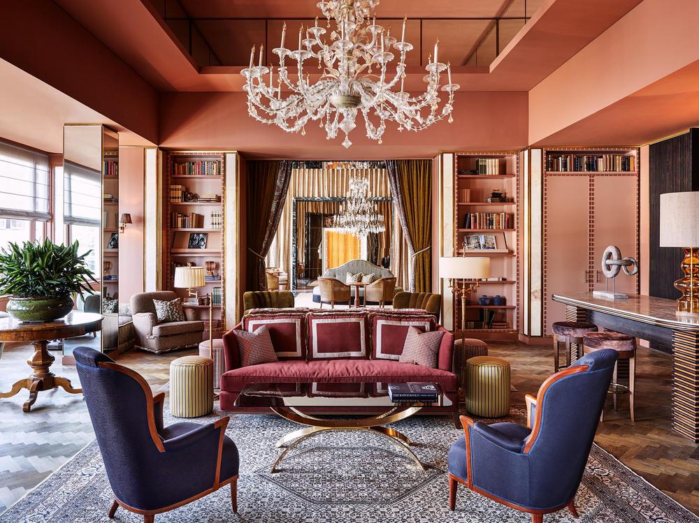 De L'Europe Amsterdam pink lounge area with a large chandelier and different furniture placed around the room with full bookcases.