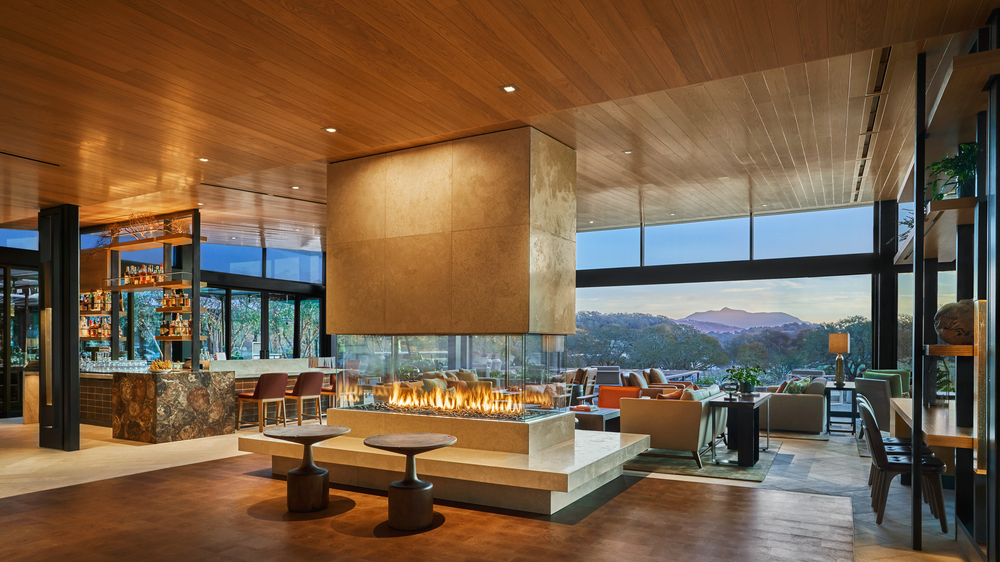 Lobby of Montage Healdsburg with a modern gold fireplace with a variety of furniture arranged in sitting area with an open air patio with beautiful views of surrounding mountains.