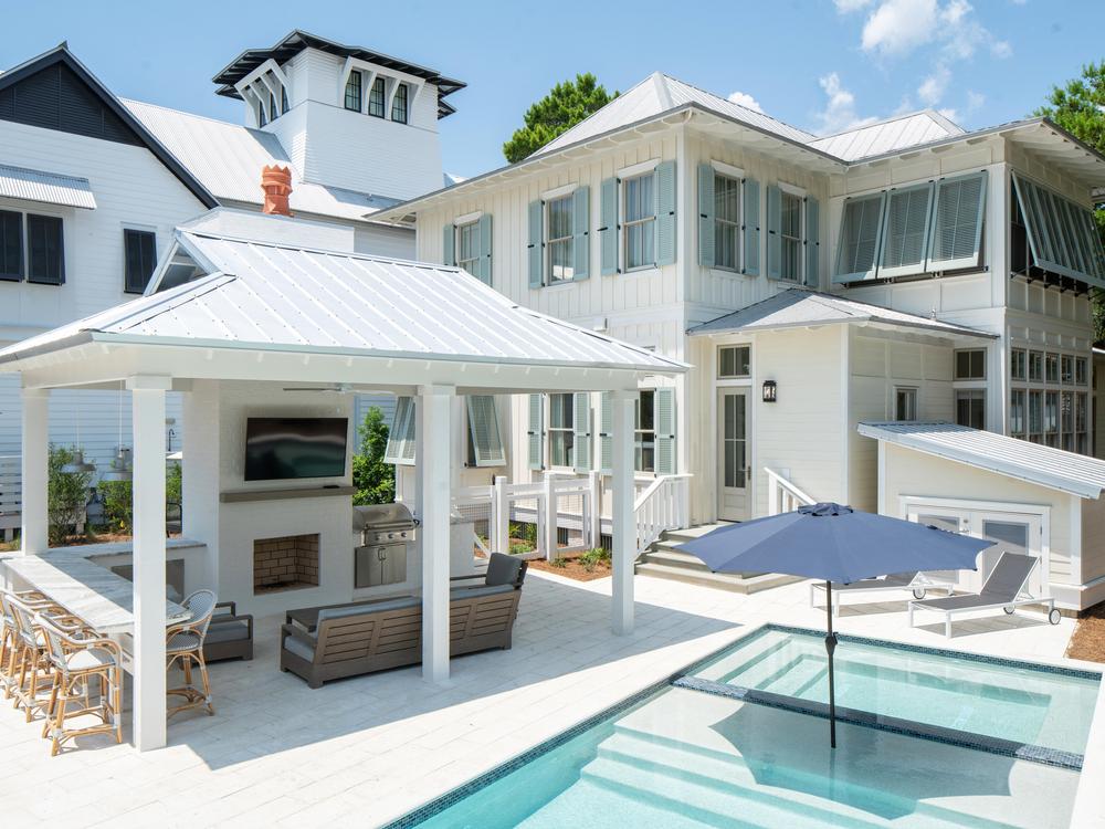 Pool area of Magnolia home with covered outdoor entertainment with a fireplace and flat screen tv with patio furniture and a large dining area. 