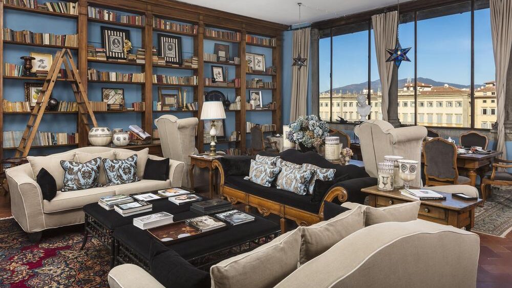 Living area with floor to ceiling bookcases with a large dining table and furniture arranged around a black coffee tables with views of Florence. 