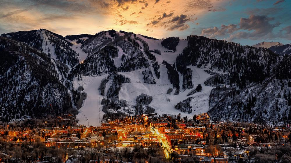 The town of Aspen illuminated during a sunset with Aspen Mountain and a light orange sky.