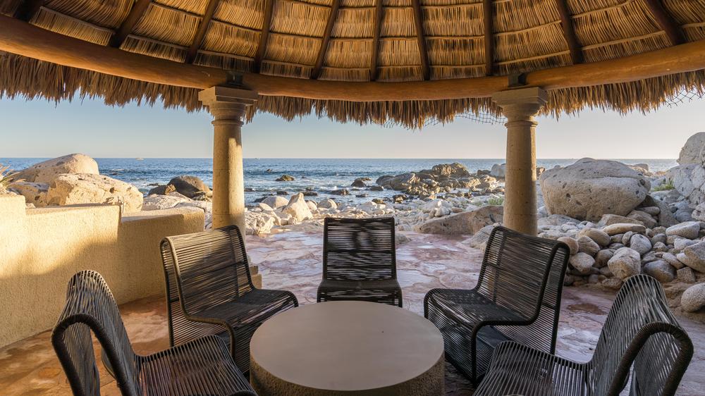 Covered palapa with a rocky beach behind it with chairs arranged around a coffee table.